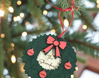 Puede incluir: Un adorno de corona navideña de madera verde con un lazo y una cinta rojos. El adorno tiene el apellido "The Sheridans" y los nombres "Andrew", "Wesley" y "Meaghan". El adorno cuelga de un árbol de Navidad.