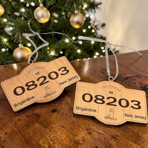 Może przedstawiać: Dwie jasne drewniane ozdoby w kształcie tablic rejestracyjnych ze srebrnymi wstążkami. Każda zawiera napis 'CHRISTMAS 2026', numer '08203', 'Brigantine' i 'New Jersey' oraz małą wygrawerowaną ilustrację latarni morskiej.