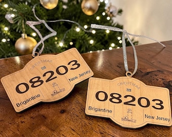 May include: Two light-colored wooden ornaments shaped like license plates with silver hanging ribbons. Each features the text 'CHRISTMAS 2026', the number '08203', 'Brigantine', and 'New Jersey', along with a small engraved lighthouse illustration.