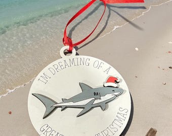 Op de afbeelding: Ronde kerstversiering met een haai met een kerstmuts. De tekst "I'M DREAMING OF A GREAT WHITE CHRISTMAS" staat in een cirkel. Een rood lint is bevestigd om op te hangen.