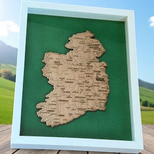 May include: A framed wooden map of Ireland, showcasing the country's counties. The map is laser-cut from light-colored wood and set against a green background within a white frame. The counties are labeled with their names.