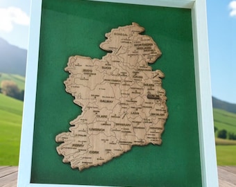 May include: A framed wooden map of Ireland, showcasing the country's counties. The map is laser-cut from light-colored wood and set against a green background within a white frame. The counties are labeled with their names.