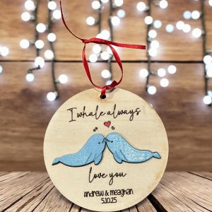 Może przedstawiać: Okrągła drewniana ozdoba z napisem "I whale always love you" oraz imionami "Andrew & Meaghan" z datą "5.10.25". Ozdoba przedstawia dwie niebieskie ilustracje wielorybów całujących się, z czerwoną wstążką do zawieszenia.