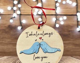 Puede incluir: Adorno redondo de madera con el texto "I whale always love you" y los nombres "Andrew & Meaghan" con la fecha "5.10.25". Presenta dos ilustraciones de ballenas azules besándose, con una cinta roja para colgar.