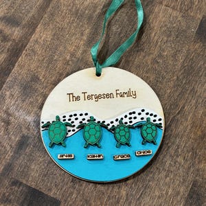 May include: A wooden family ornament with the text "The Tergesen Family". The ornament features four green turtles above names: Arlie, Kathin, Grace, and Chloe, set against a blue and white background, with a green ribbon.