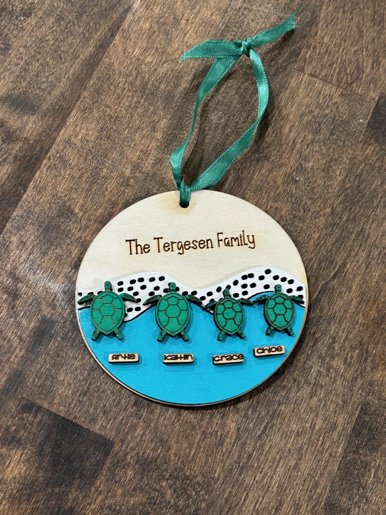 May include: A wooden family ornament with the text "The Tergesen Family". The ornament features four green turtles above names: Arlie, Kathin, Grace, and Chloe, set against a blue and white background, with a green ribbon.