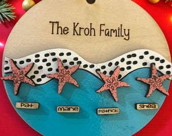 Puede incluir: Un letrero familiar redondo de madera con el texto "The Kroh Family". El letrero presenta un diseño de océano azul con cuatro estrellas de mar rosas y etiquetas de nombres individuales. Hay un agujero en la parte superior para colgar.