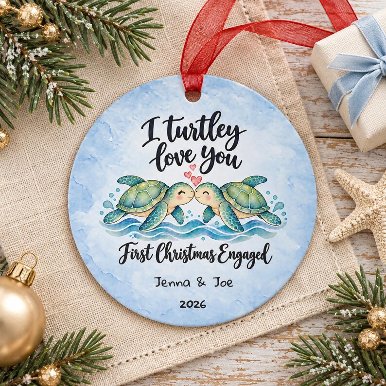 May include: A round ceramic Christmas ornament with a light blue watercolour background featuring two illustrated sea turtles touching noses, three small red hearts, and the text I turtley love you, First Christmas Engaged, Jenna & Joe, 2026.