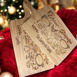 May include: Two wooden serving boards with handles, engraved with holiday-themed text and designs. The boards feature text that reads "Dear Santa" and "Love, The Santorufo Family". Designs include a reindeer, Christmas tree, and cookie.