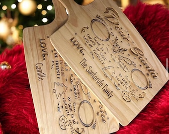 May include: Two wooden serving boards with handles, engraved with holiday-themed text and designs. The boards feature text that reads "Dear Santa" and "Love, The Santorufo Family". Designs include a reindeer, Christmas tree, and cookie.