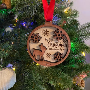 Op de afbeelding: Een houten kerstbal met een herten silhouet, sneeuwvlokken en de tekst "My First Christmas Mackenzie Grace 2022". De ornament hangt aan een rood lint in een kerstboom.