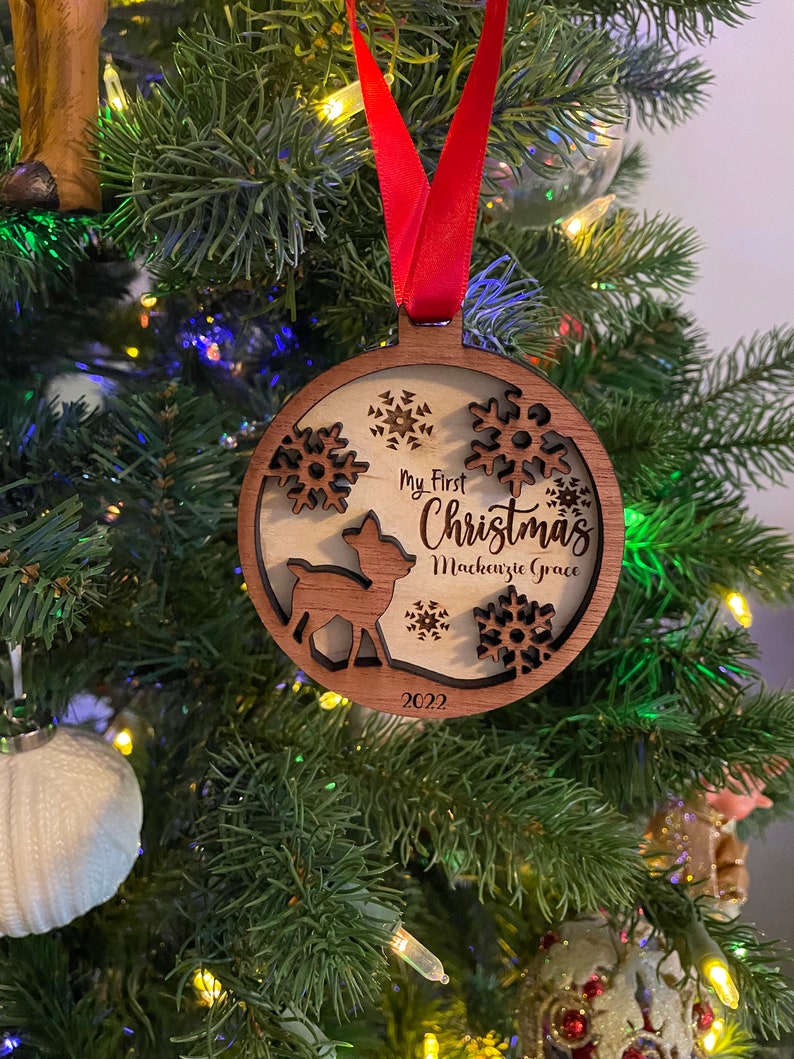 Op de afbeelding: Een houten kerstbal met een herten silhouet, sneeuwvlokken en de tekst "My First Christmas Mackenzie Grace 2022". De ornament hangt aan een rood lint in een kerstboom.