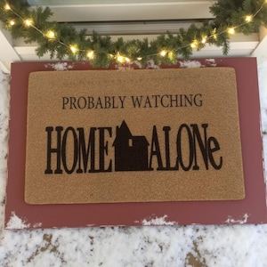 May include: A rectangular doormat with the text "PROBABLY WATCHING HOME ALONE" in black. The mat is a light brown color and is set in a red frame. A string of lights and greenery are above the mat.