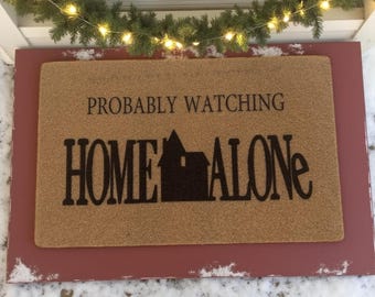 May include: A rectangular doormat with the text "PROBABLY WATCHING HOME ALONE" in black. The mat is a light brown color and is set in a red frame. A string of lights and greenery are above the mat.