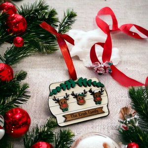 May include: A wooden Christmas ornament featuring three reindeer with red noses and green bow ties. The ornament has a banner that reads "The Kroh Family" and is decorated with green leaves. A red ribbon is attached for hanging.