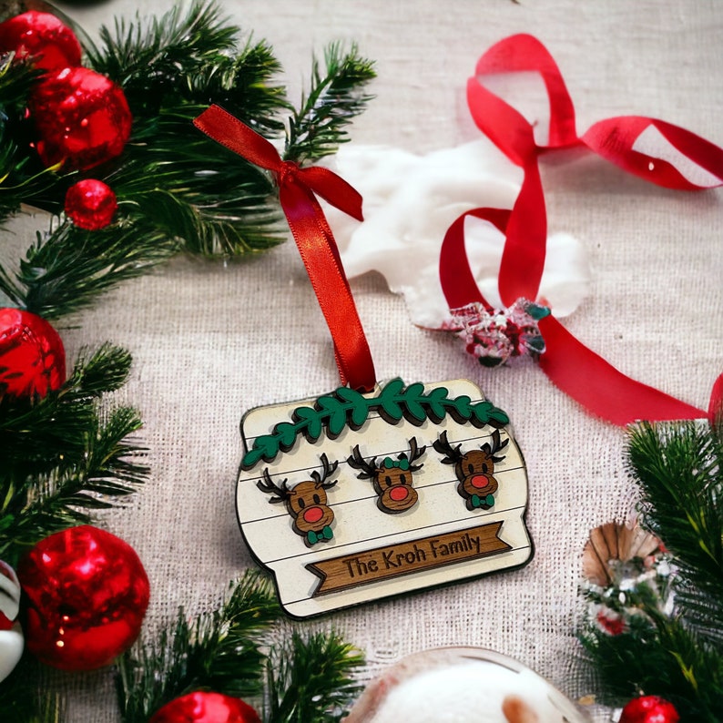 May include: A wooden Christmas ornament featuring three reindeer with red noses and green bow ties. The ornament has a banner that reads "The Kroh Family" and is decorated with green leaves. A red ribbon is attached for hanging.