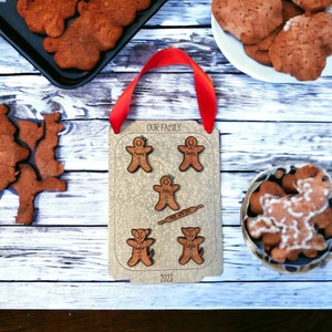 Puede incluir: Un adorno familiar de madera con figuras de pan de jengibre y perros, el texto "OUR FAMILY" y "2023". El adorno tiene una cinta roja para colgar. Galletas de jengibre se ven en el fondo.