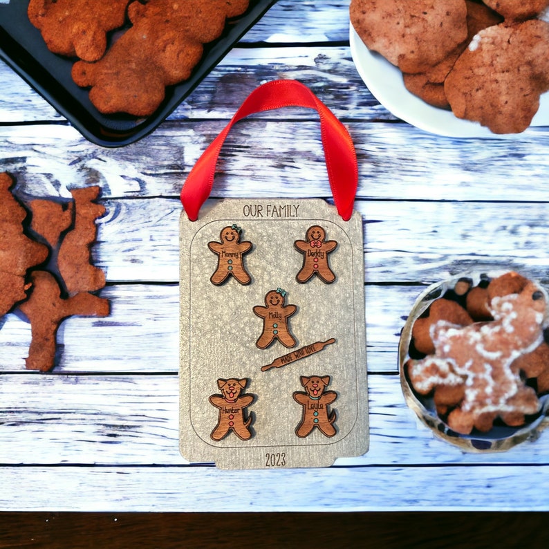 Puede incluir: Un adorno familiar de madera con figuras de pan de jengibre y perros, el texto "OUR FAMILY" y "2023". El adorno tiene una cinta roja para colgar. Galletas de jengibre se ven en el fondo.