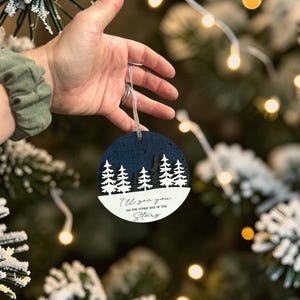 May include: A circular layered ornament featuring a dark blue night sky with cut-out stars above a forest of white and dark grey pine trees. The white base displays the black text: I'll see you ON THE OTHER SIDE OF THE Stars. It hangs by a silver cord.