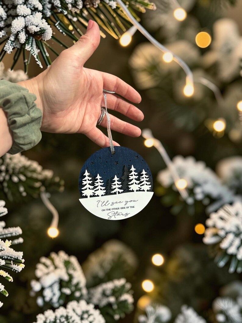 May include: A circular layered ornament featuring a dark blue night sky with cut-out stars above a forest of white and dark grey pine trees. The white base displays the black text: I'll see you ON THE OTHER SIDE OF THE Stars. It hangs by a silver cord.