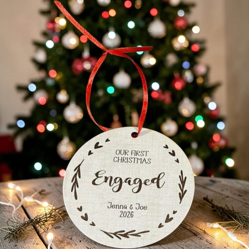 May include: A round, light-coloured wooden ornament featuring a red ribbon loop. The text reads "OUR FIRST CHRISTMAS Engaged" with the names "Jenna & Joe" and the year "2026" engraved in black, surrounded by a decorative border of small hearts and leaves.