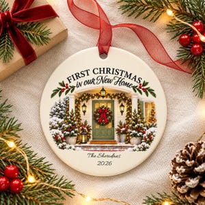 May include: A round white ceramic ornament featuring a printed illustration of a house entrance decorated for the holidays. The text reads First Christmas in our New Home, The Sheridans, and 2026. A red ribbon is attached to the top for hanging.