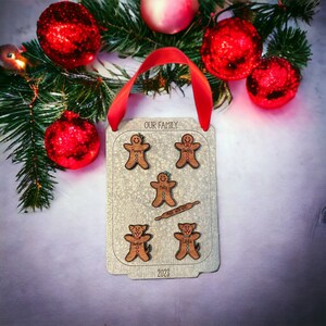 Puede incluir: Adorno navide&ntilde;o personalizado con una cinta roja, con figuras de hombrecitos de jengibre y perros. El adorno dice "OUR FAMILY" y "Made with Love", con nombres y el a&ntilde;o 2023. Decoraciones navide&ntilde;as rodean el adorno.