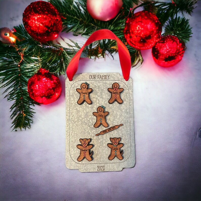 Puede incluir: Adorno navide&ntilde;o personalizado con una cinta roja, con figuras de hombrecitos de jengibre y perros. El adorno dice "OUR FAMILY" y "Made with Love", con nombres y el a&ntilde;o 2023. Decoraciones navide&ntilde;as rodean el adorno.