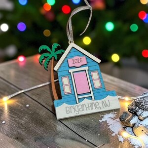 Puede incluir: Un adorno de madera con forma de caseta de playa azul, puerta rosa y palmera verde. El adorno muestra el texto 2026 sobre la puerta y Brigantine, NJ en la base. Incluye una cinta plateada para colgarlo en el &aacute;rbol de Navidad.