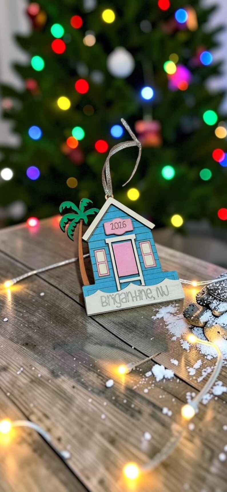 Puede incluir: Un adorno de madera con forma de caseta de playa azul, puerta rosa y palmera verde. El adorno muestra el texto 2026 sobre la puerta y Brigantine, NJ en la base. Incluye una cinta plateada para colgarlo en el &aacute;rbol de Navidad.