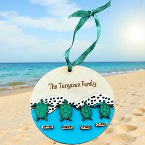 May include: A wooden family ornament with the text "The Tergesen Family". The ornament features four green sea turtles above the names Arha, Kahn, Grace, and Chloe. It has a teal ribbon for hanging.