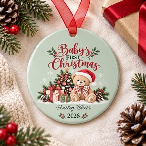 May include: A round, light green ceramic ornament with a red ribbon hanger. The design features a teddy bear wearing a Santa hat, a decorated Christmas tree, and gifts. Text reads Baby's First Christmas, Hailey Blair, and the year 2026.