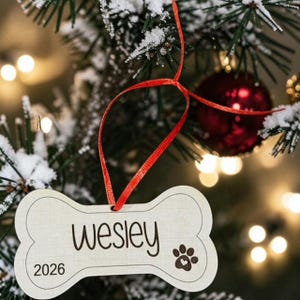 May include: A bone-shaped light-colored wooden ornament hanging from a red ribbon. The ornament features the name Wesley in dark script, the year 2026, and a paw print icon with a small heart inside the center pad, all printed in a dark color.