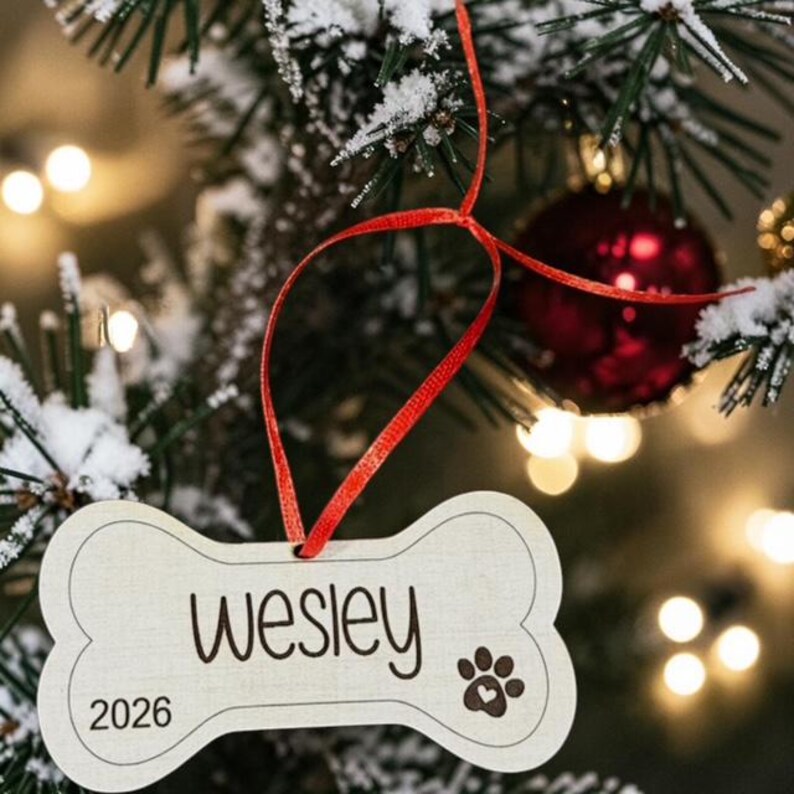 May include: A bone-shaped light-colored wooden ornament hanging from a red ribbon. The ornament features the name Wesley in dark script, the year 2026, and a paw print icon with a small heart inside the center pad, all printed in a dark color.