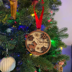 Op de afbeelding: Een houten kerstversiering met een rood lint, hangend aan een kerstboom. De versiering heeft een herten silhouet, sneeuwvlokken en de tekst "My First Christmas Mackenzie Grace 2022".