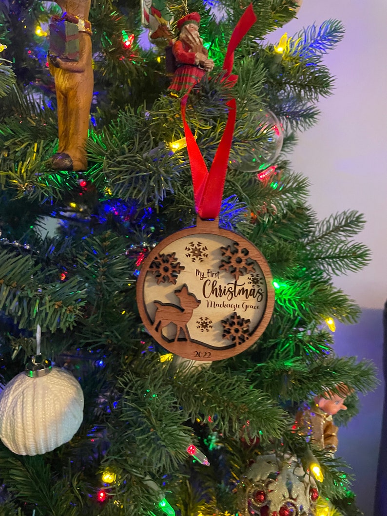 Op de afbeelding: Een houten kerstversiering met een rood lint, hangend aan een kerstboom. De versiering heeft een herten silhouet, sneeuwvlokken en de tekst "My First Christmas Mackenzie Grace 2022".