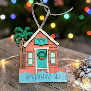 Puede incluir: Un adorno de madera con forma de caseta de playa en color coral, con puerta turquesa, corona verde y una palmera. El texto 2026 aparece sobre la puerta y la base tiene la inscripci&oacute;n Brigantine, NJ. Incluye una cinta plateada para colgarlo.
