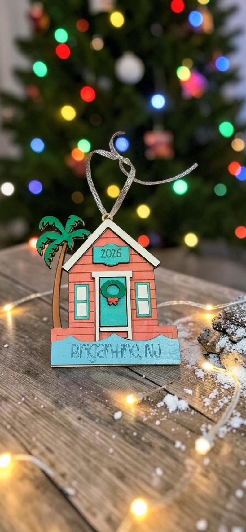 Puede incluir: Un adorno de madera con forma de caseta de playa en color coral, con puerta turquesa, corona verde y una palmera. El texto 2026 aparece sobre la puerta y la base tiene la inscripci&oacute;n Brigantine, NJ. Incluye una cinta plateada para colgarlo.