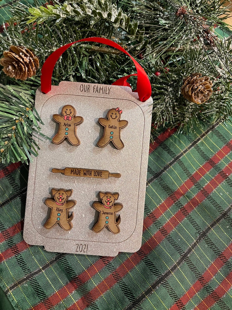 Puede incluir: Adorno familiar de madera con figuras de hombre de jengibre y perros. El adorno dice "OUR FAMILY" y "MADE WITH LOVE". Tambi&eacute;n se muestra el a&ntilde;o "2021". Se adjunta una cinta roja para colgar.