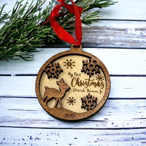Może przedstawiać: Drewniana ozdoba choinkowa z sylwetką jelenia, płatkami śniegu i napisem "My First Christmas Patrick Thomas Jr. 2023." Ozdoba ma czerwoną wstążkę do zawieszenia i jest wykonana z warstwowego drewna.