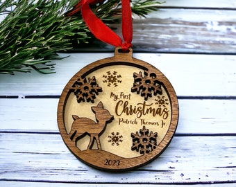 Puede incluir: Adorno navideño de madera con un ciervo, copos de nieve y el texto "My First Christmas Patrick Thomas Jr. 2023." Tiene una cinta roja y está hecho de madera en capas. Decoración festiva.