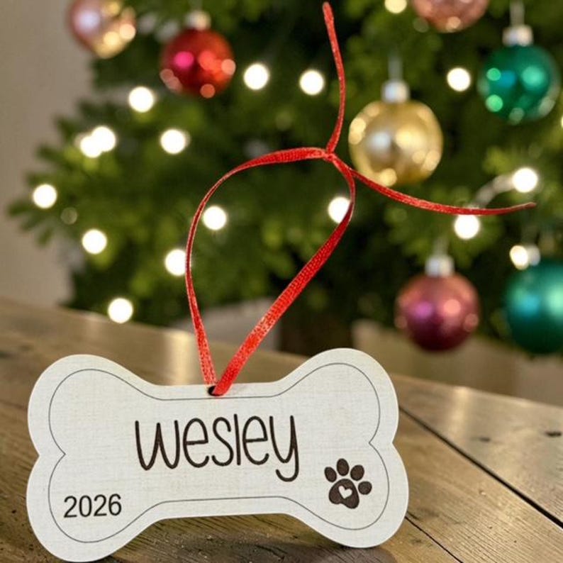 May include: A bone-shaped wooden ornament featuring the name Wesley printed in black script, the year 2026, and a paw print icon with a small heart in the center. A thin red ribbon is attached to the top for hanging the light-colored wooden piece.