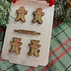 Puede incluir: Adorno familiar de madera con forma de bandeja de horno, con figuras de hombrecitos de jengibre y perros. El adorno tiene el texto "OUR FAMILY", "MADE WITH LOVE" y "2021". Est&aacute; decorado con una cinta roja para colgar.