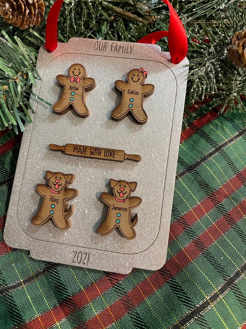 Puede incluir: Adorno familiar de madera con forma de bandeja de horno, con figuras de hombrecitos de jengibre y perros. El adorno tiene el texto "OUR FAMILY", "MADE WITH LOVE" y "2021". Est&aacute; decorado con una cinta roja para colgar.