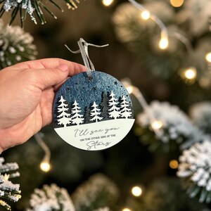 May include: A round, flat, wooden Christmas ornament featuring a dark blue, glittery night sky with cut-out stars above a layered forest of white and black pine trees. The bottom white section displays the text, I'll see you on the other side of the stars.
