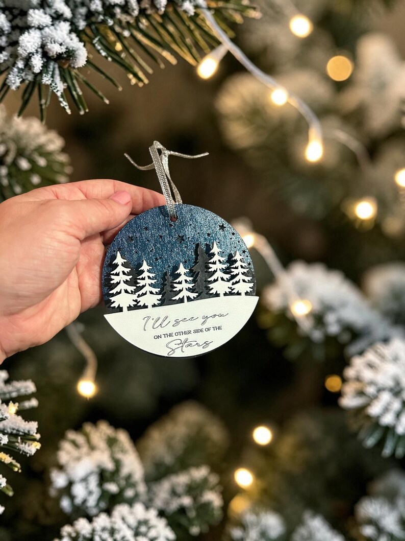 May include: A round, flat, wooden Christmas ornament featuring a dark blue, glittery night sky with cut-out stars above a layered forest of white and black pine trees. The bottom white section displays the text, I'll see you on the other side of the stars.
