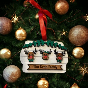 May include: A wooden Christmas ornament featuring three reindeer with red noses and green bow ties, above the text "The Kroh Family." The ornament is white with a wooden border and green leaf accents, hanging from a red ribbon.