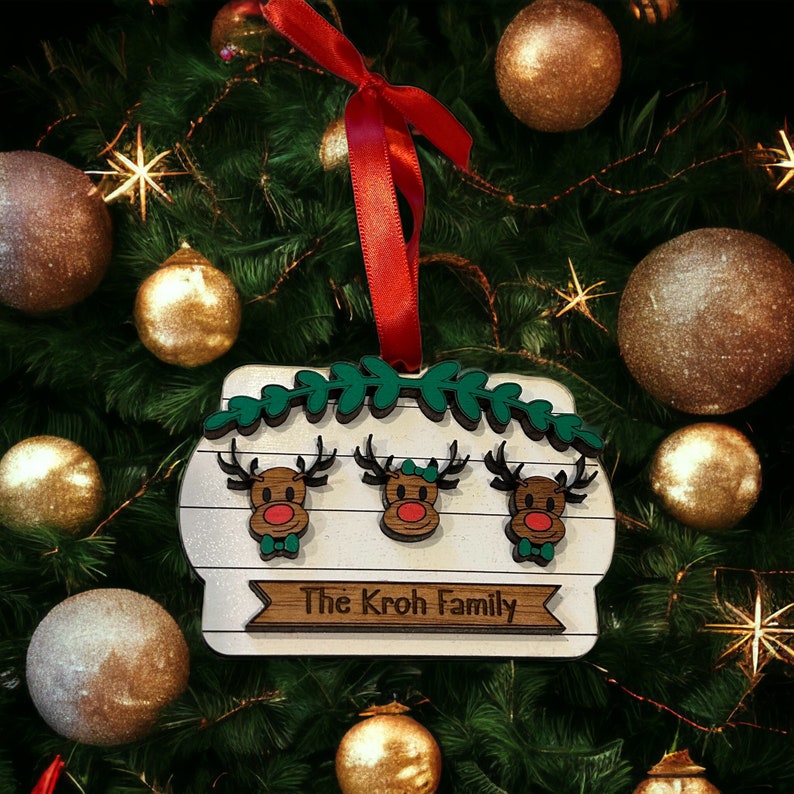 May include: A wooden Christmas ornament featuring three reindeer with red noses and green bow ties, above the text "The Kroh Family." The ornament is white with a wooden border and green leaf accents, hanging from a red ribbon.