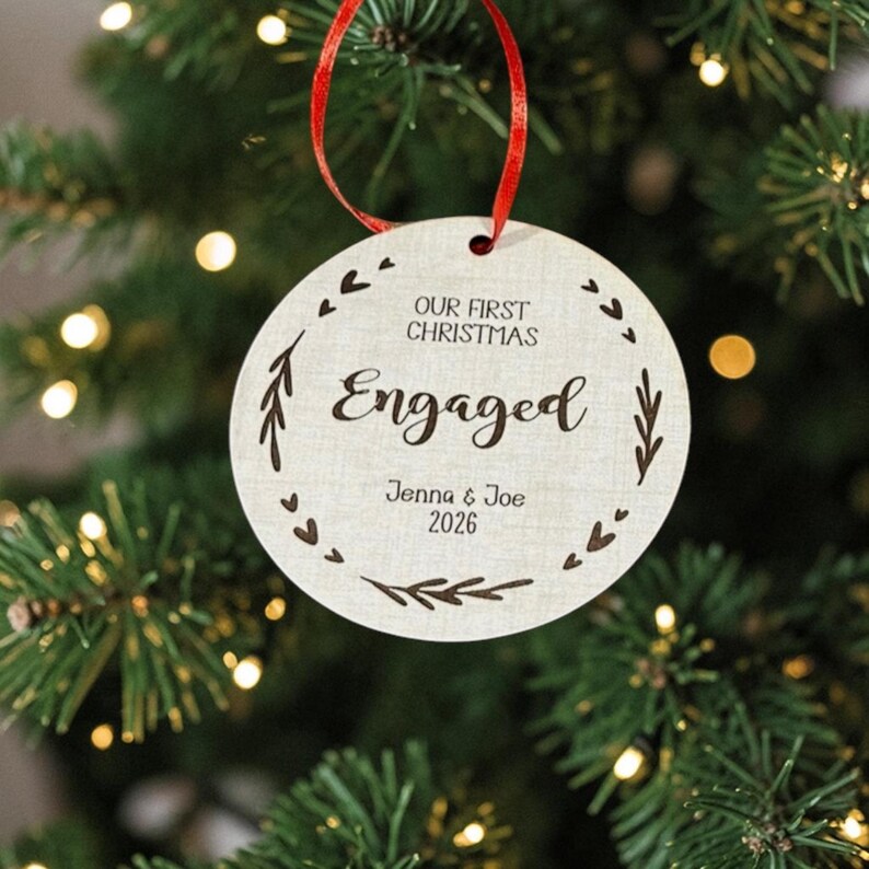 May include: A round, light-coloured wooden ornament hanging from a red ribbon. The text reads "OUR FIRST CHRISTMAS", "Engaged", "Jenna & Joe", and "2026". The design features a circular border of small etched hearts and leafy branch motifs.