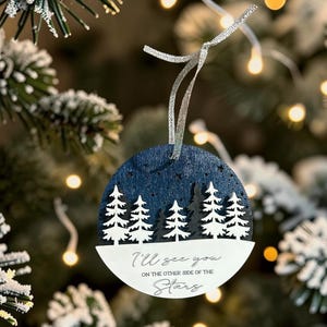 May include: A circular wooden ornament featuring a dark blue night sky with a forest of white pine trees. The lower white section displays the black text: I'll see you ON THE OTHER SIDE OF THE Stars. A silver ribbon is attached to the top for hanging.